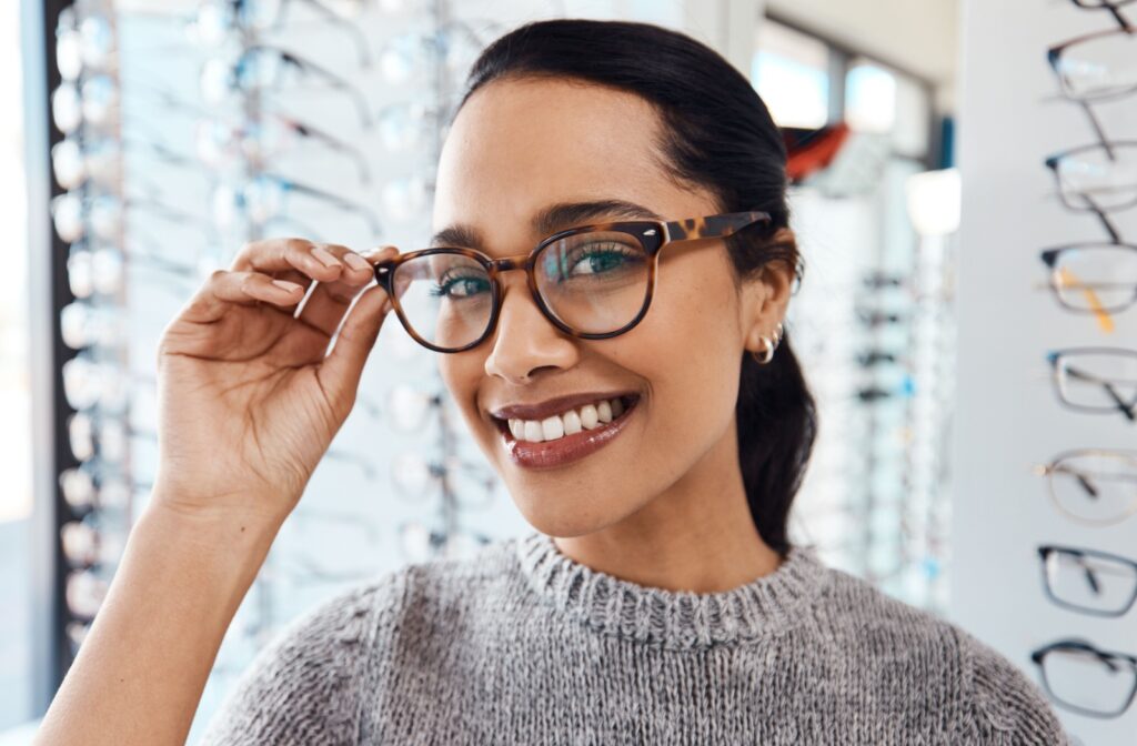 A woman looking into the camera with a wide smile and her left hand on her new pair of eyeglasses.