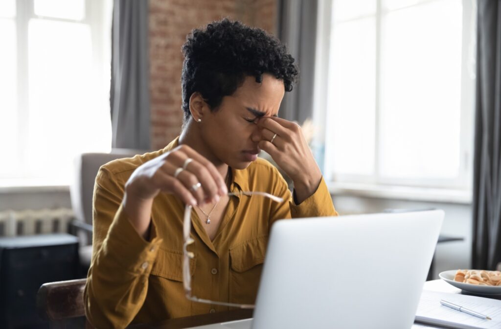 An adult working from home removes their glasses to pinch the bridge of their nose in an attempt to stop an oncoming headache