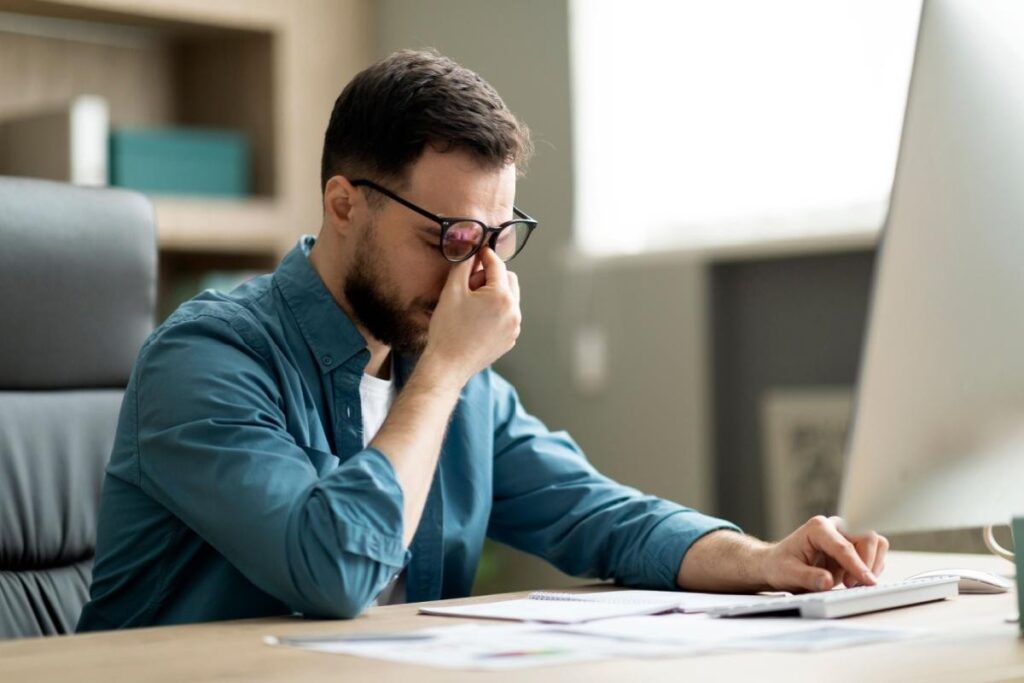 A professional man wearing glasses sits at an office desk, looking stressed and tired while rubbing the bridge of his nose to alleviate eye fatigue caused by prolonged computer screen use.