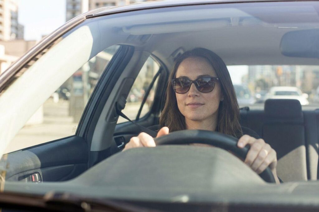 Adult wearing sunglasses driving a car in an urban setting, both hands on the steering wheel, viewed through the windshield.